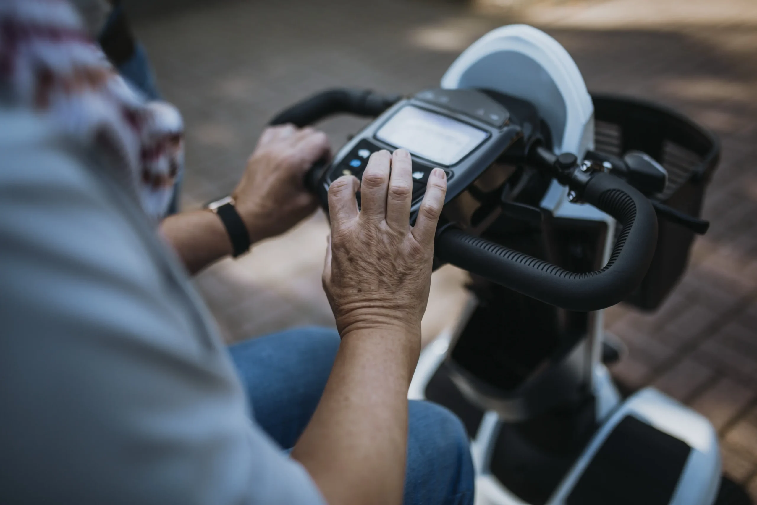 Ältere Person bedient die Steuerung eines Elektromobils, Nahaufnahme der Hände am Lenker mit Display, Sitz und Fußablage des Fahrzeugs sichtbar, draußen auf gepflastertem Weg.