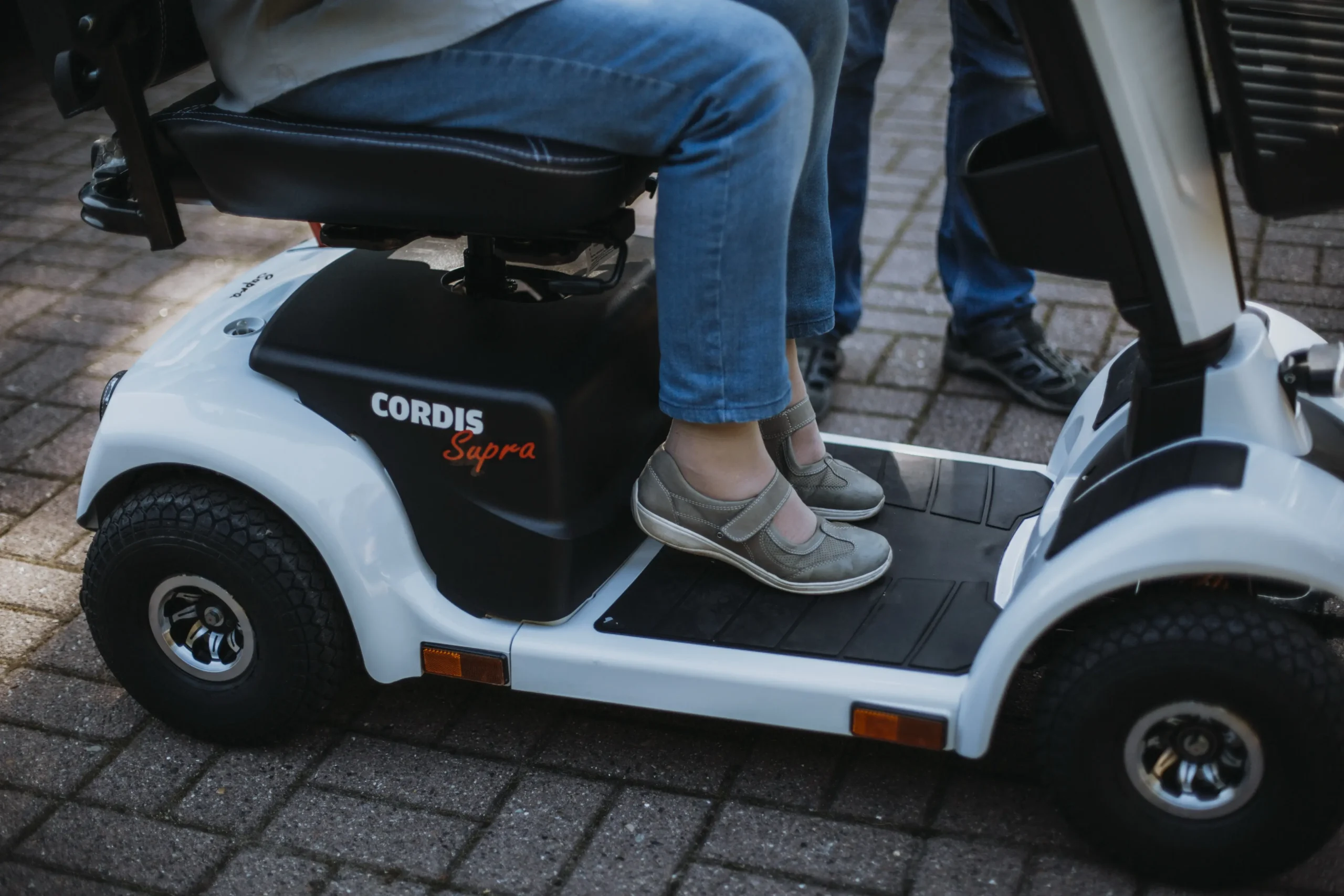 Person sitzt auf einem weißen Elektromobil der Marke Cordis Supra, zu sehen sind die Beine mit blauen Jeans und grauen Schuhen. Das Elektromobil steht auf gepflastertem Untergrund, im Hintergrund sind weitere Beine von Personen erkennbar.