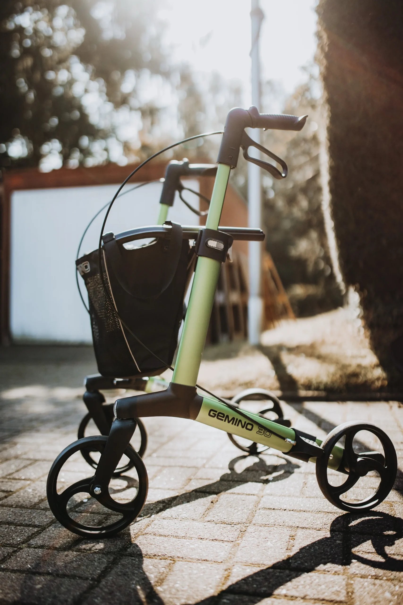Leichtgrüner, dreirädriger Rollator mit schwarzem Griff, Handbremse und Einkaufstasche, steht auf gepflastertem Weg im Freien, Modellbezeichnung „GEMINO 30“ sichtbar.