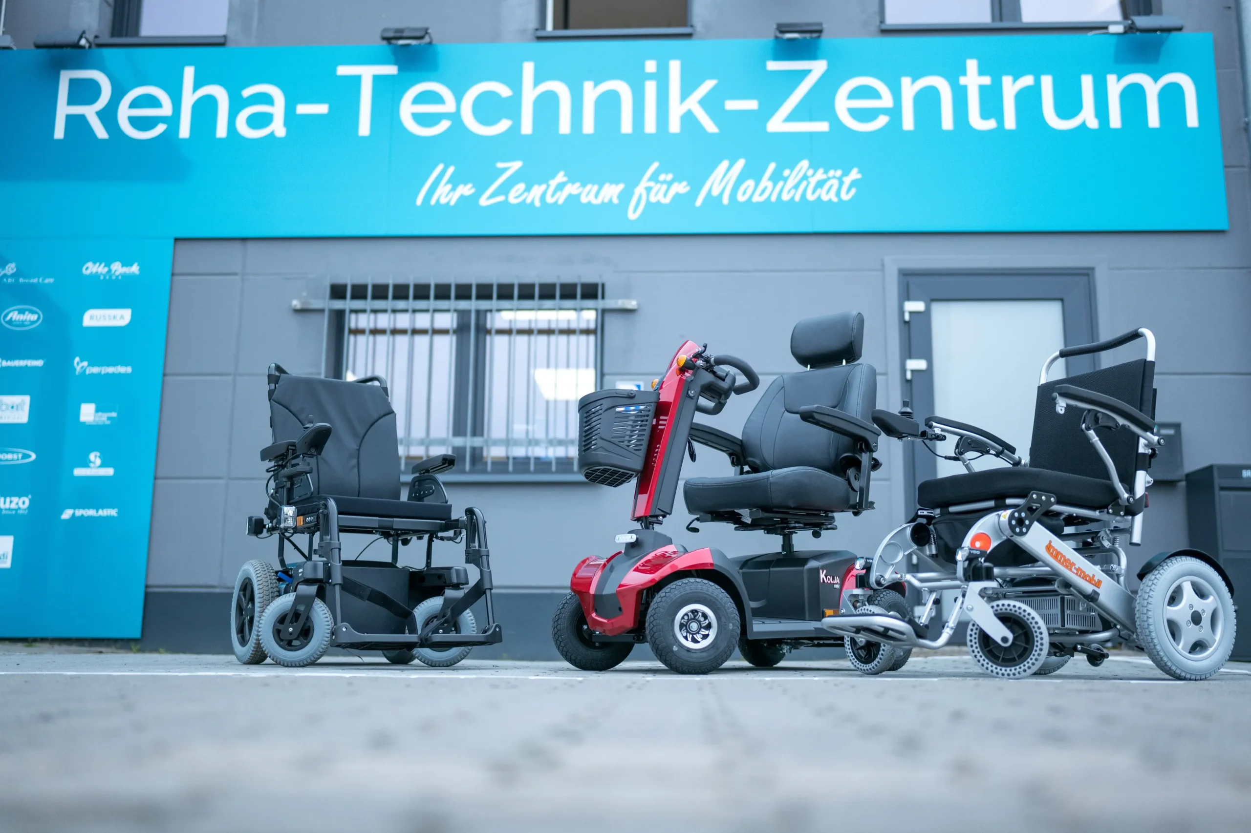 Drei verschiedene Mobilitätshilfen vor einem Gebäude mit großem Schild „Reha-Technik-Zentrum Ihr Zentrum für Mobilität“: ein schwarzer Elektrorollstuhl links, ein rotes Elektromobil in der Mitte und ein weiterer schwarzer Elektrorollstuhl rechts. Das Gebäude ist grau, links neben dem Eingang ist eine blaue Tafel mit Logos verschiedener Marken.