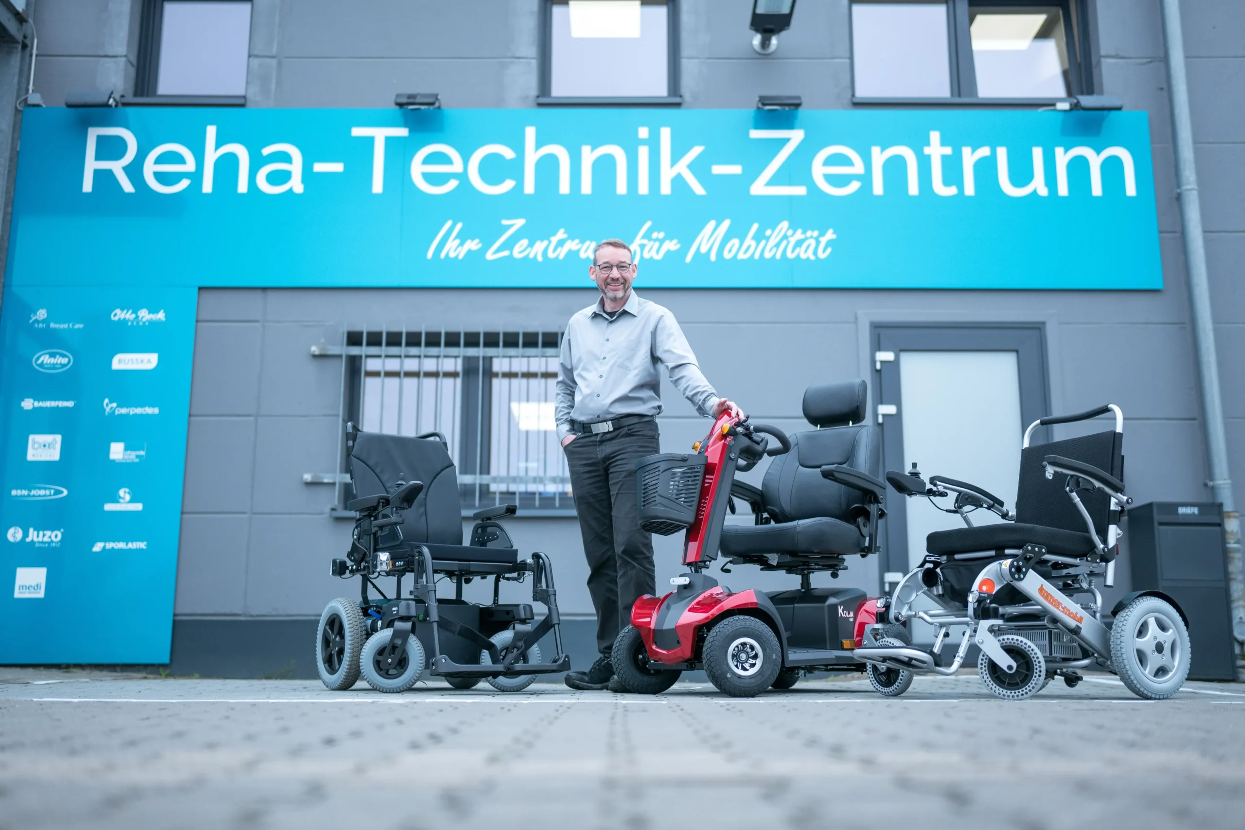 Mann steht vor dem Reha-Technik-Zentrum mit drei Mobilitätshilfen: zwei elektrische Rollstühle und ein Elektromobil in Rot. Gebäude mit großem blauen Schild „Reha-Technik-Zentrum Ihr Zentrum für Mobilität“ und Logos verschiedener Hersteller an der Wand. Szene vermittelt Beratung und Angebot von Rehatechnik und Mobilitätshilfen.