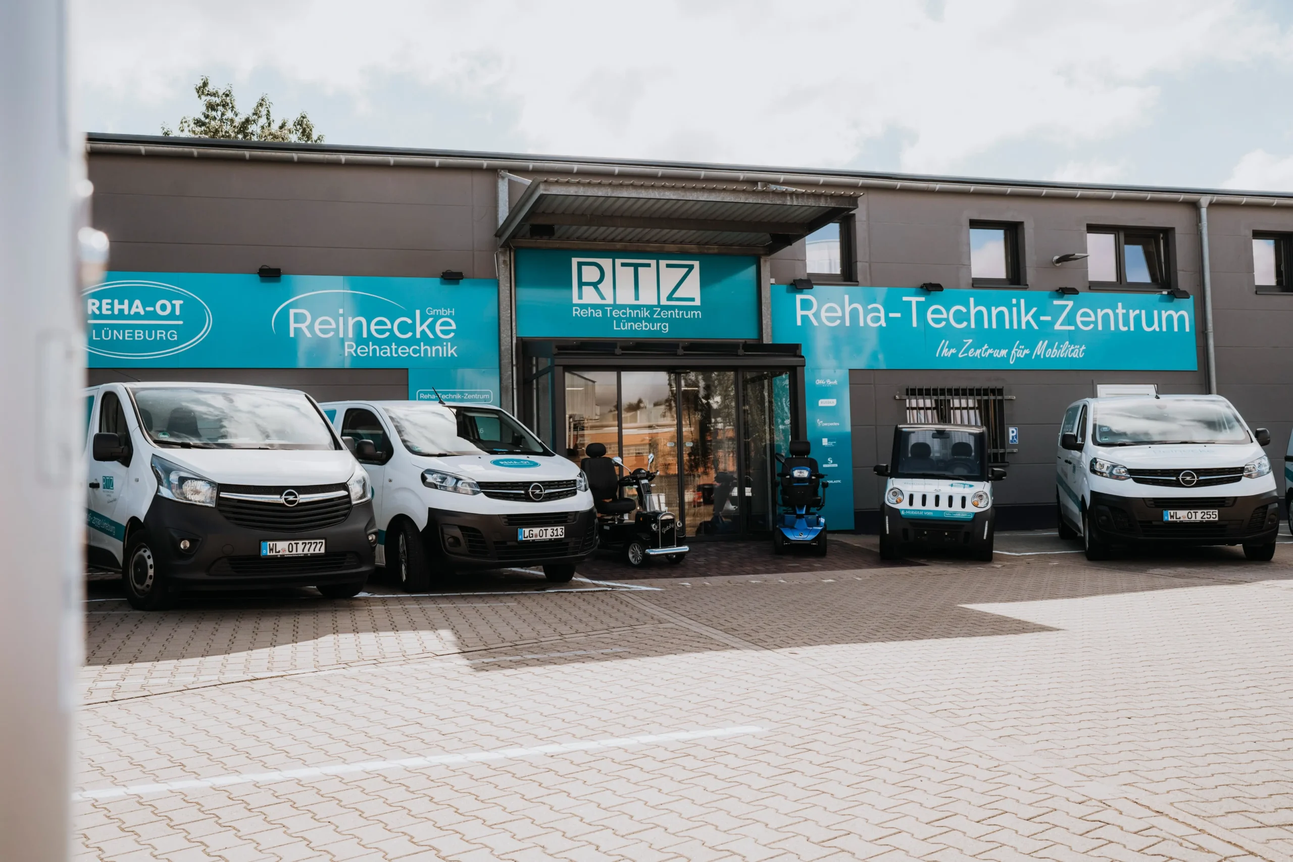 Eingangsbereich des Reha-Technik-Zentrums Lüneburg mit drei weißen Firmenfahrzeugen und verschiedenen Mobilitätshilfen wie Elektrorollstühlen und Rollstühlen vor dem Gebäude. Große blaue Schilder mit den Aufschriften „RTZ Reha Technik Zentrum Lüneburg“, „Reinecke Rehatechnik“ und „Reha-Technik-Zentrum Ihr Zentrum für Mobilität“ sind gut sichtbar. Parkplatz mit gepflastertem Boden vor dem Gebäude.