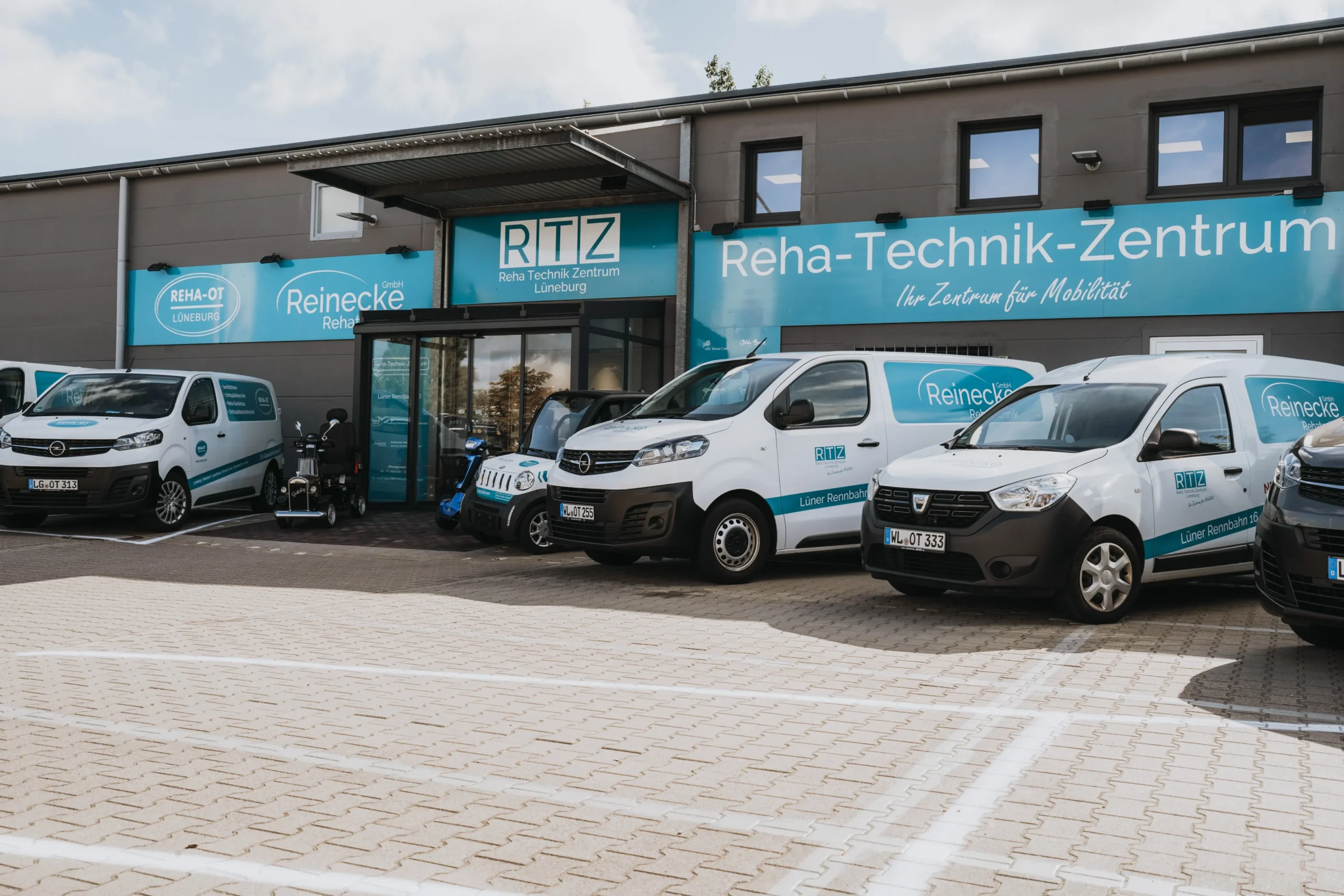 Außenansicht des Reha-Technik-Zentrums Lüneburg mit mehreren weißen Firmenfahrzeugen, die mit dem Logo von Rehatechnik Reinecke und dem Schriftzug „Lüner Rehazentrum“ beklebt sind. Große blaue Fassadenbeschilderung mit der Aufschrift „Reha-Technik-Zentrum Ihr Zentrum für Mobilität“ sowie weitere Logos von Rehatechnik Reinecke und REHA-OT Lüneburg. Parkplatz mit gepflasterter Fläche vor dem Gebäude.