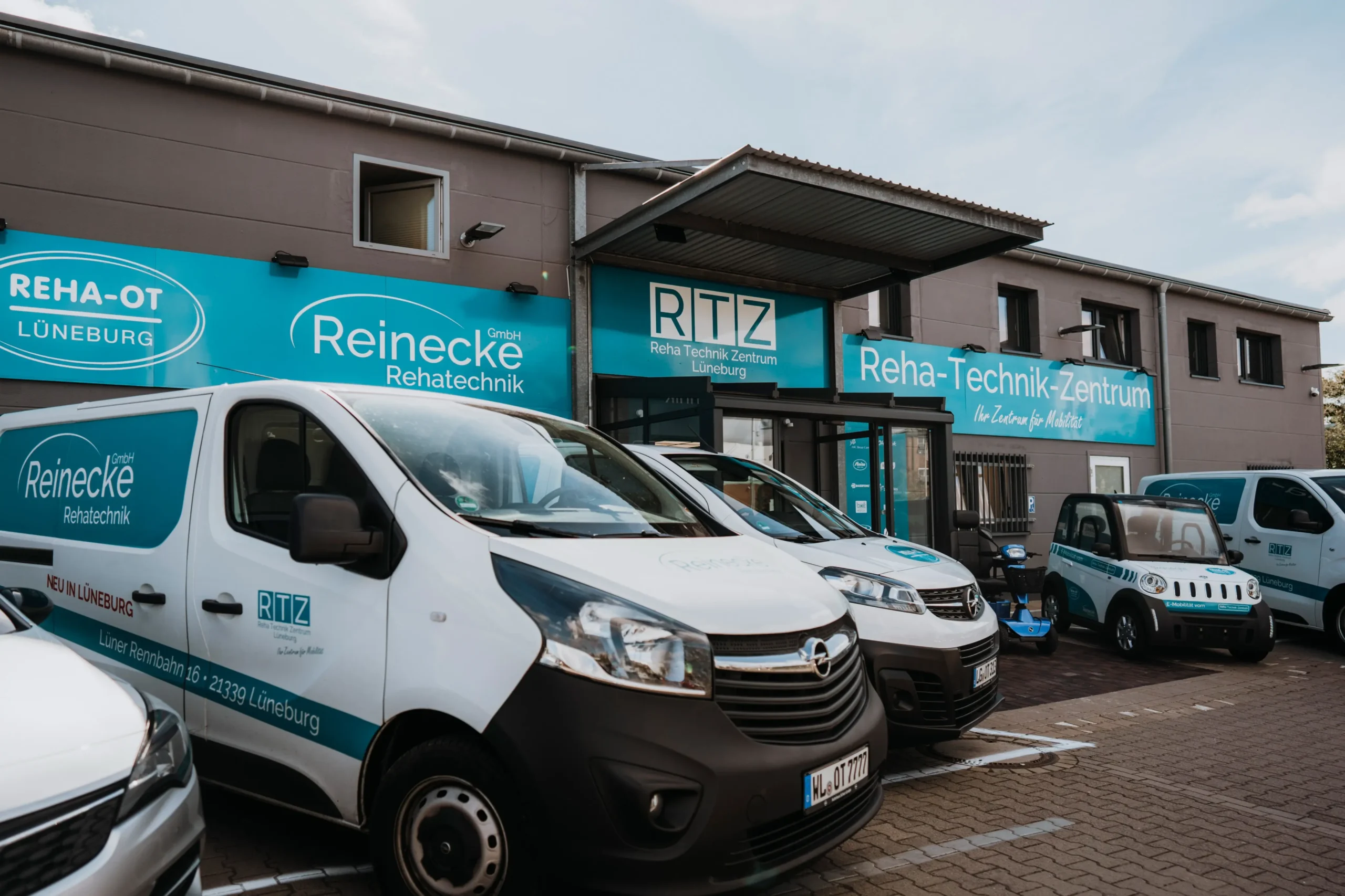 Außenansicht des Reha-Technik-Zentrums Lüneburg mit mehreren weißen Firmenfahrzeugen, darunter Transporter mit dem Schriftzug „Reinecke Rehatechnik GmbH“ und „RTZ Reha Technik Zentrum Lüneburg“. Gebäude mit blauen Firmenschildern, die Rehatechnik und Reha-OT Lüneburg bewerben. Parkplatz mit mehreren Fahrzeugen vor dem Eingang.