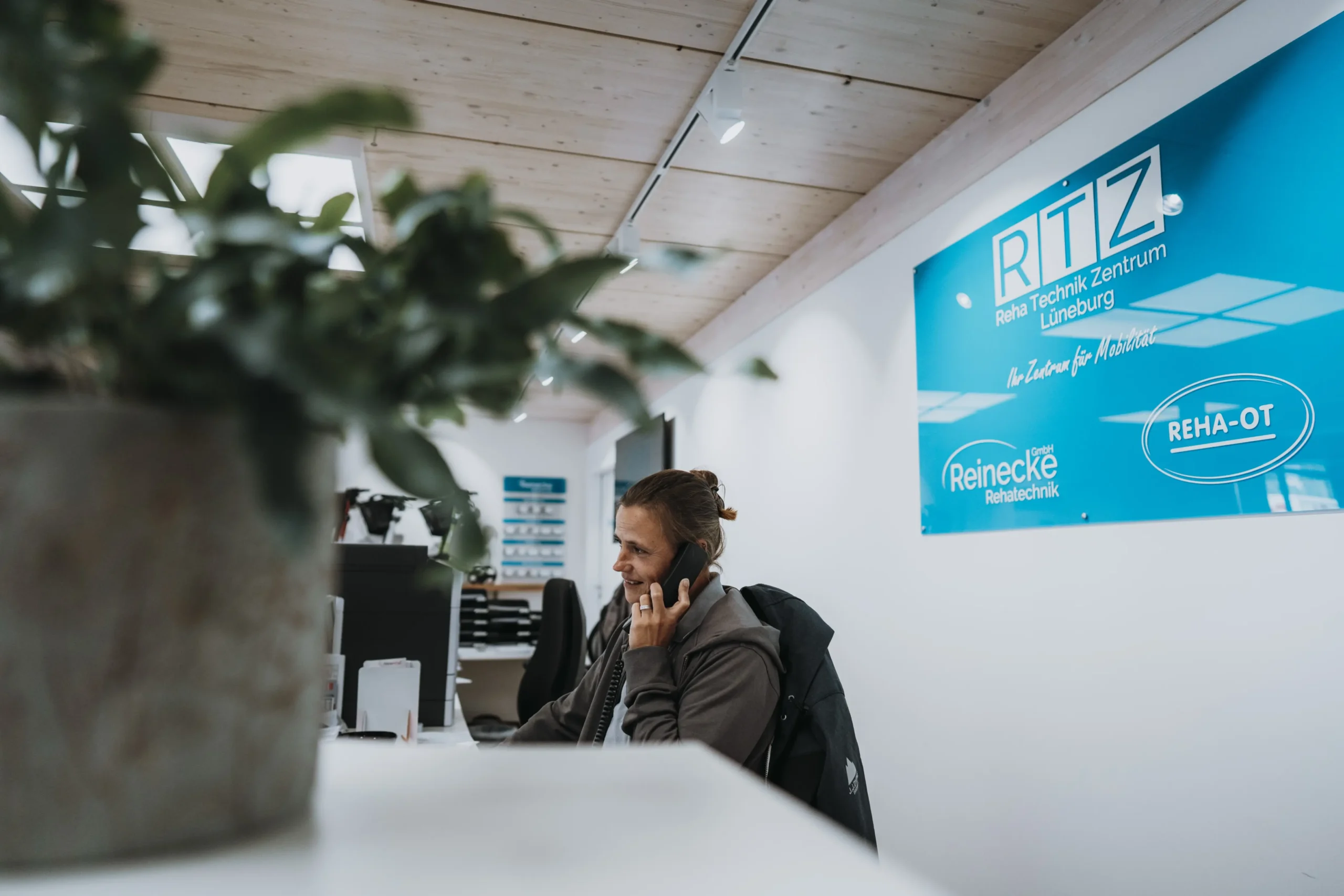 Mitarbeiterin am Empfangstresen eines Sanitätshauses, telefoniert und lächelt, im Hintergrund Wandtafel mit den Logos „RTZ Reha Technik Zentrum Lüneburg“, „Reinecke Rehatechnik“ und „REHA-OT“, helle, moderne Innenraumgestaltung.