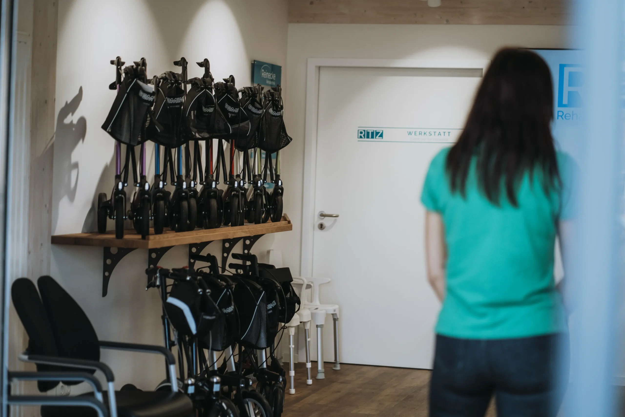 Mehrere zusammengeklappte Rollatoren stehen auf zwei Regalen an einer Wand, daneben sind weiße Gehhilfen zu sehen. Im Vordergrund steht eine Person mit dunklen Haaren und grünem Oberteil, die auf eine weiße Tür mit dem Schild „RTZ WERKSTATT“ blickt. Der Raum wirkt hell und freundlich, typisch für ein Sanitätshaus oder eine Rehatechnik-Werkstatt.