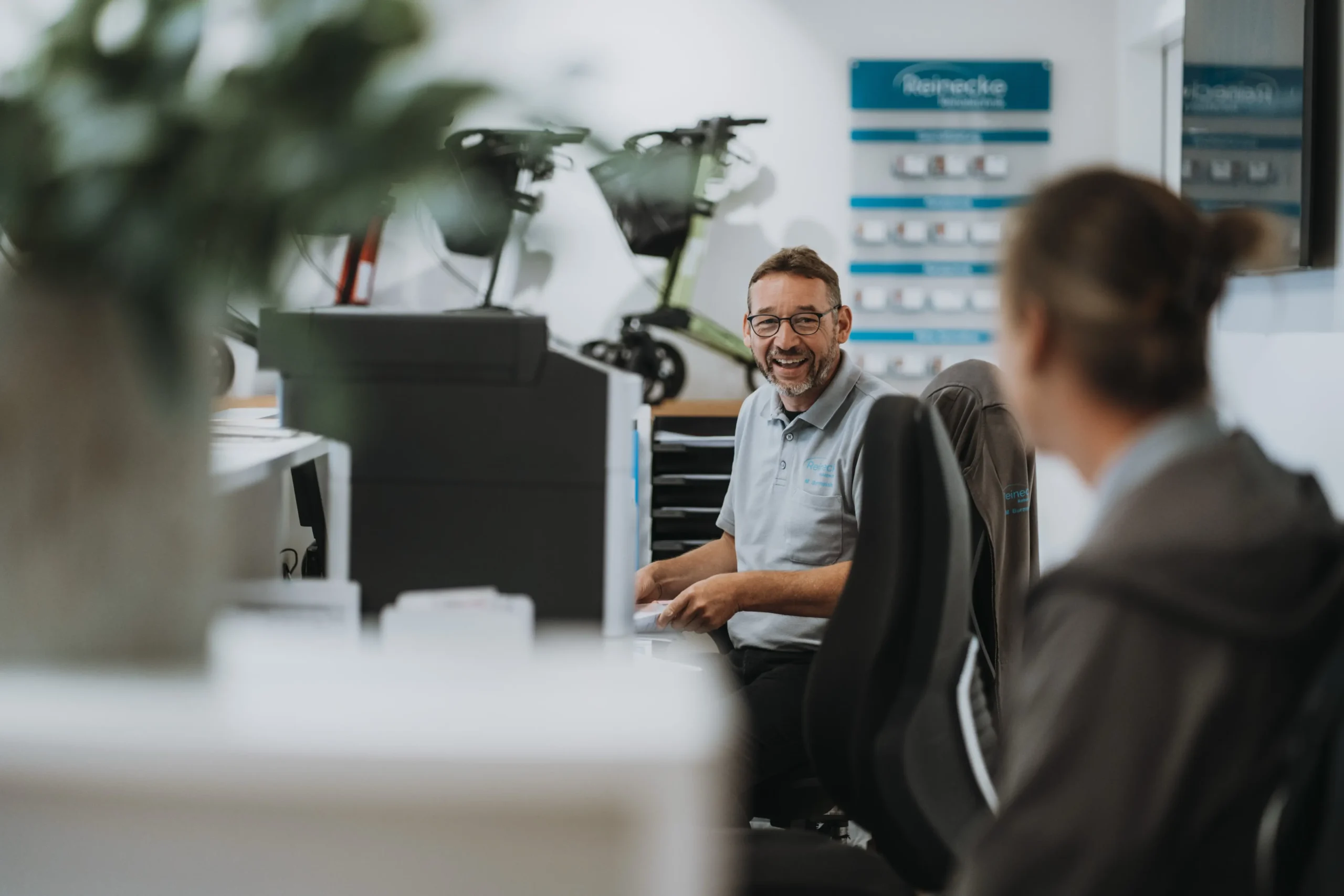 Mann mit Brille und grauem Poloshirt sitzt lächelnd an einem Schreibtisch in einem hellen Büro, im Hintergrund sind zwei Elektrorollstühle und ein Schild mit dem Schriftzug „Reinecke“ zu sehen, eine weitere Person sitzt im Vordergrund unscharf.