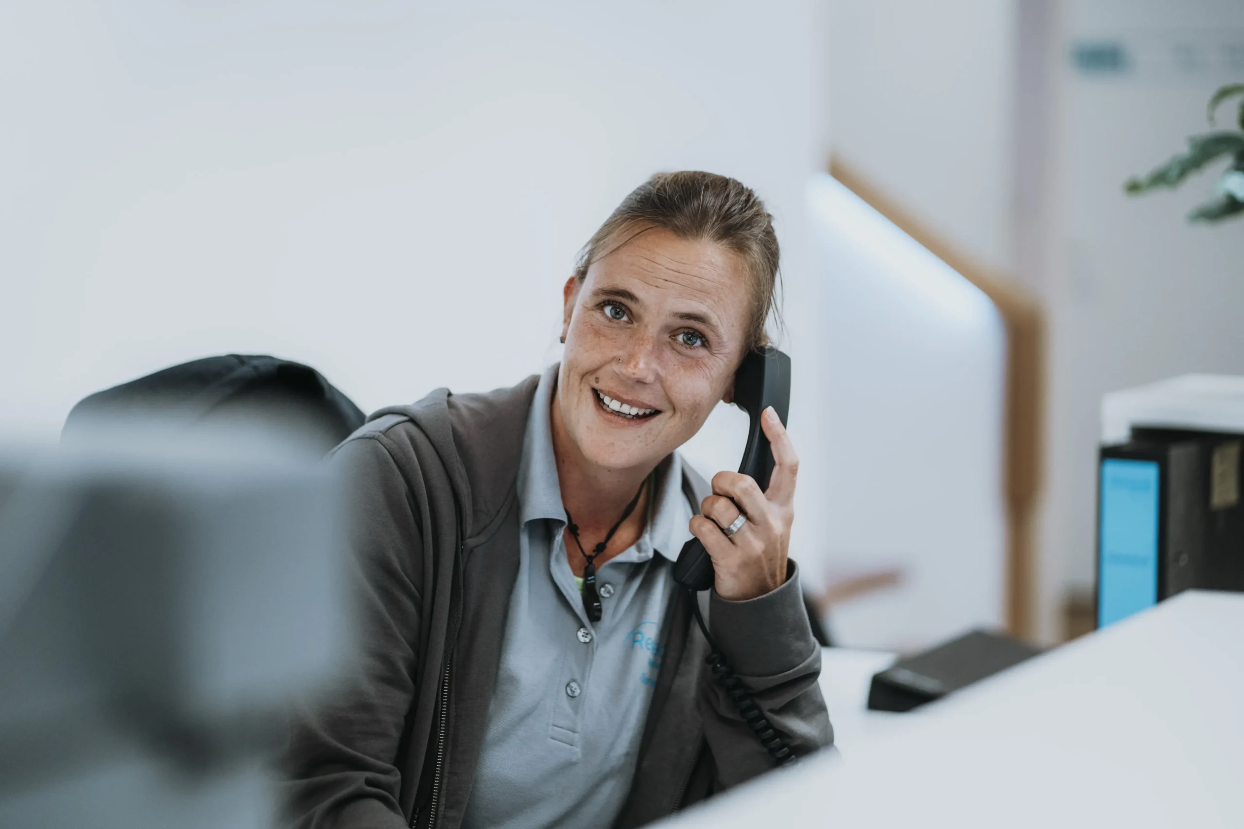Frau am Empfangstresen spricht freundlich in ein Telefon, trägt graues Poloshirt und Jacke, helle, moderne Büroatmosphäre im Hintergrund.