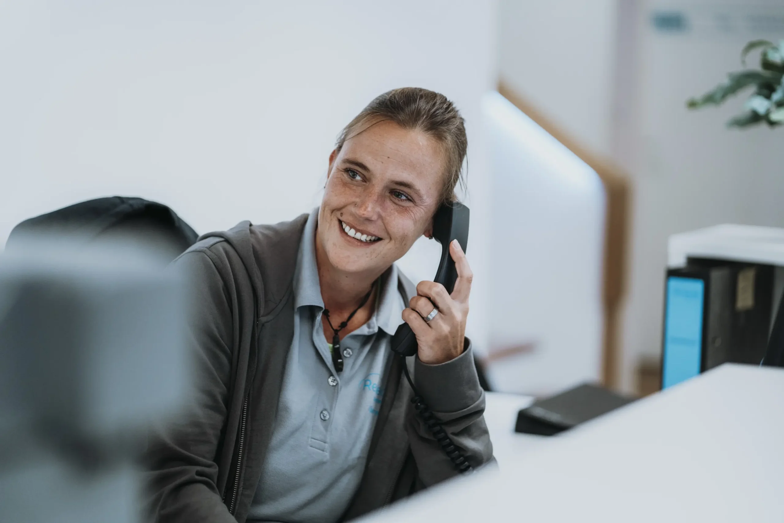 Mitarbeiterin am Empfang eines Sanitätshauses, lächelnd und telefonierend, trägt graues Poloshirt und Jacke, im Hintergrund Büroausstattung mit Monitor und Pflanzen, freundliche Atmosphäre, Rehatechnik Lüneburg und Winsen.