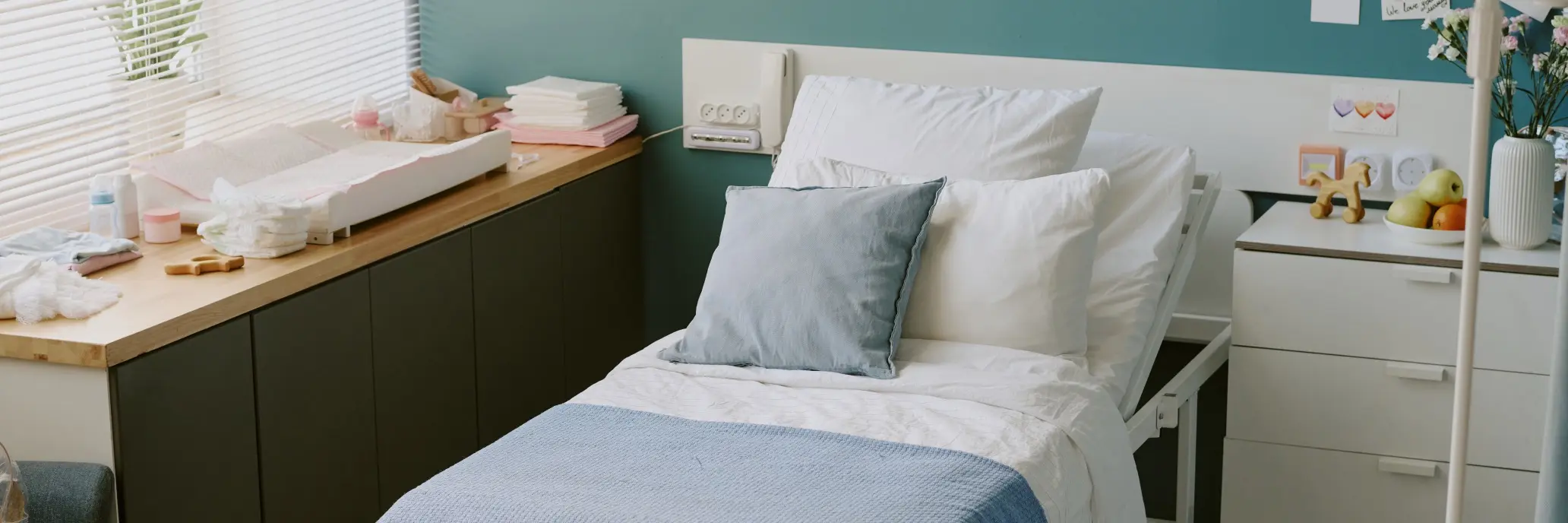Hell eingerichtetes, gemütliches Krankenzimmer mit verstellbarem Pflegebett, mehreren Kissen und sauberer Bettwäsche. Neben dem Bett steht ein Nachtschrank mit Obstschale, Vase mit Blumen und kleinen Dekorationen. Links befindet sich eine Fensterbank mit Pflegeutensilien und Handtüchern. Die Wand im Hintergrund ist zweifarbig gestaltet.