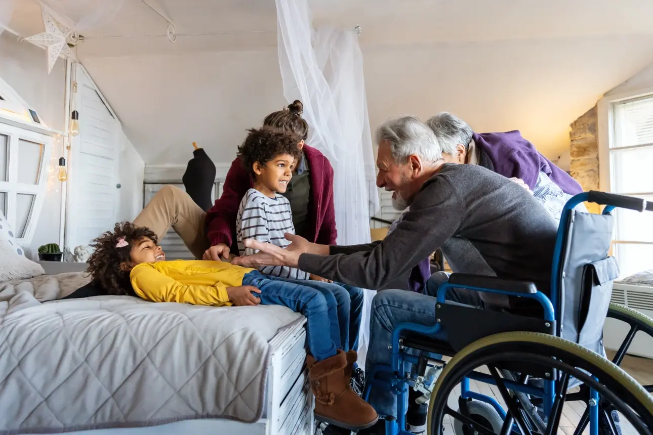 Mehrgenerationenfamilie in einem hellen Wohnzimmer, ein älterer Mann im Rollstuhl spricht freundlich mit zwei Kindern, eine Frau sitzt hinter den Kindern auf dem Bett, eine weitere ältere Person ist teilweise sichtbar, die Szene vermittelt Nähe und Fürsorge.