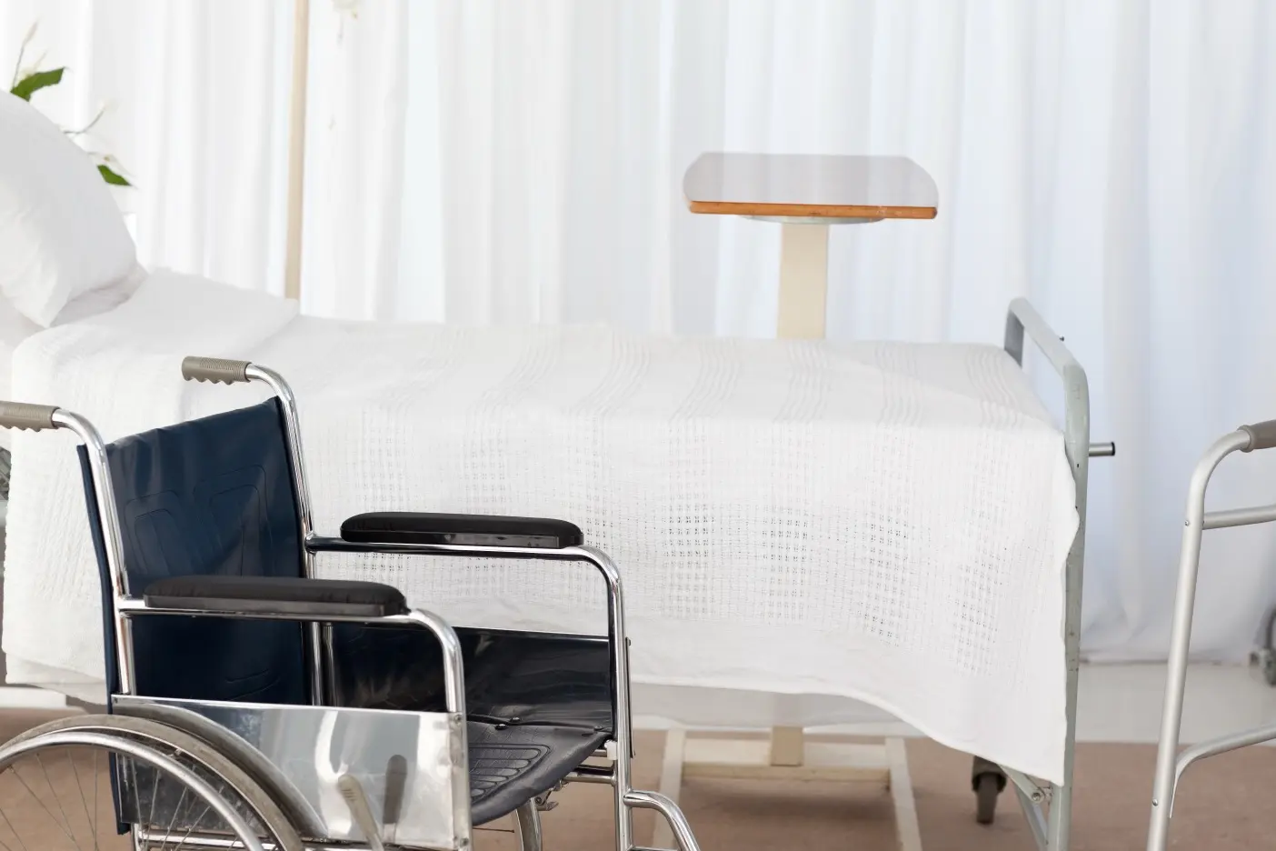 Krankenhauszimmer mit weißem Pflegebett, über dem ein höhenverstellbarer Tisch steht, davor ein schwarzer Rollstuhl mit Metallrahmen und rechts ein silberner Gehwagen. Helle, freundliche Atmosphäre mit weißen Vorhängen im Hintergrund.