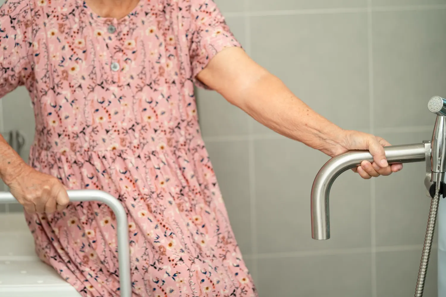 Ältere Person in rosa geblümtem Kleid hält sich mit einer Hand an einem Haltegriff im Badezimmer fest, die andere Hand greift einen weiteren Haltegriff neben einer Dusche, graue Fliesen im Hintergrund, barrierefreie Ausstattung für mehr Sicherheit und Mobilität im Bad.