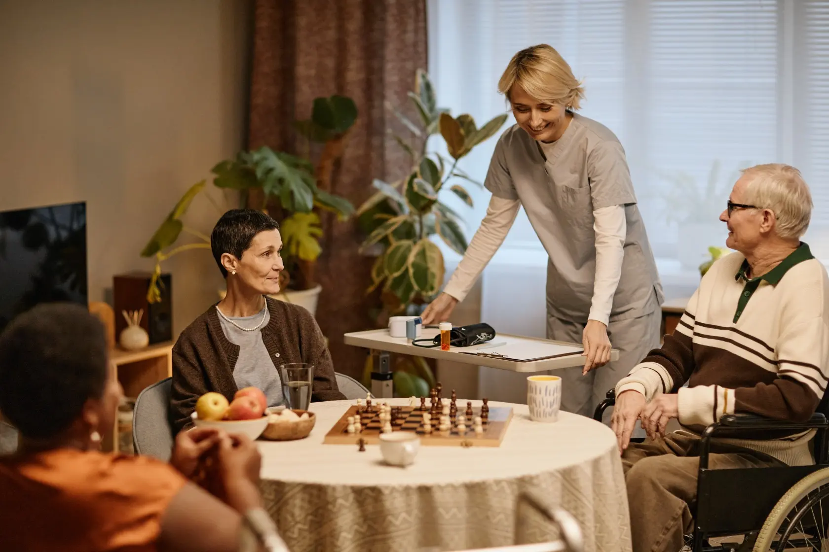 Seniorin und Senior im Rollstuhl sitzen an einem runden Tisch mit Schachspiel und Obstschale, eine Pflegerin in grauer Arbeitskleidung steht lächelnd daneben und hält ein Tablett mit Medikamenten und einem Blutdruckmessgerät, helle Wohnzimmereinrichtung mit Pflanzen im Hintergrund.
