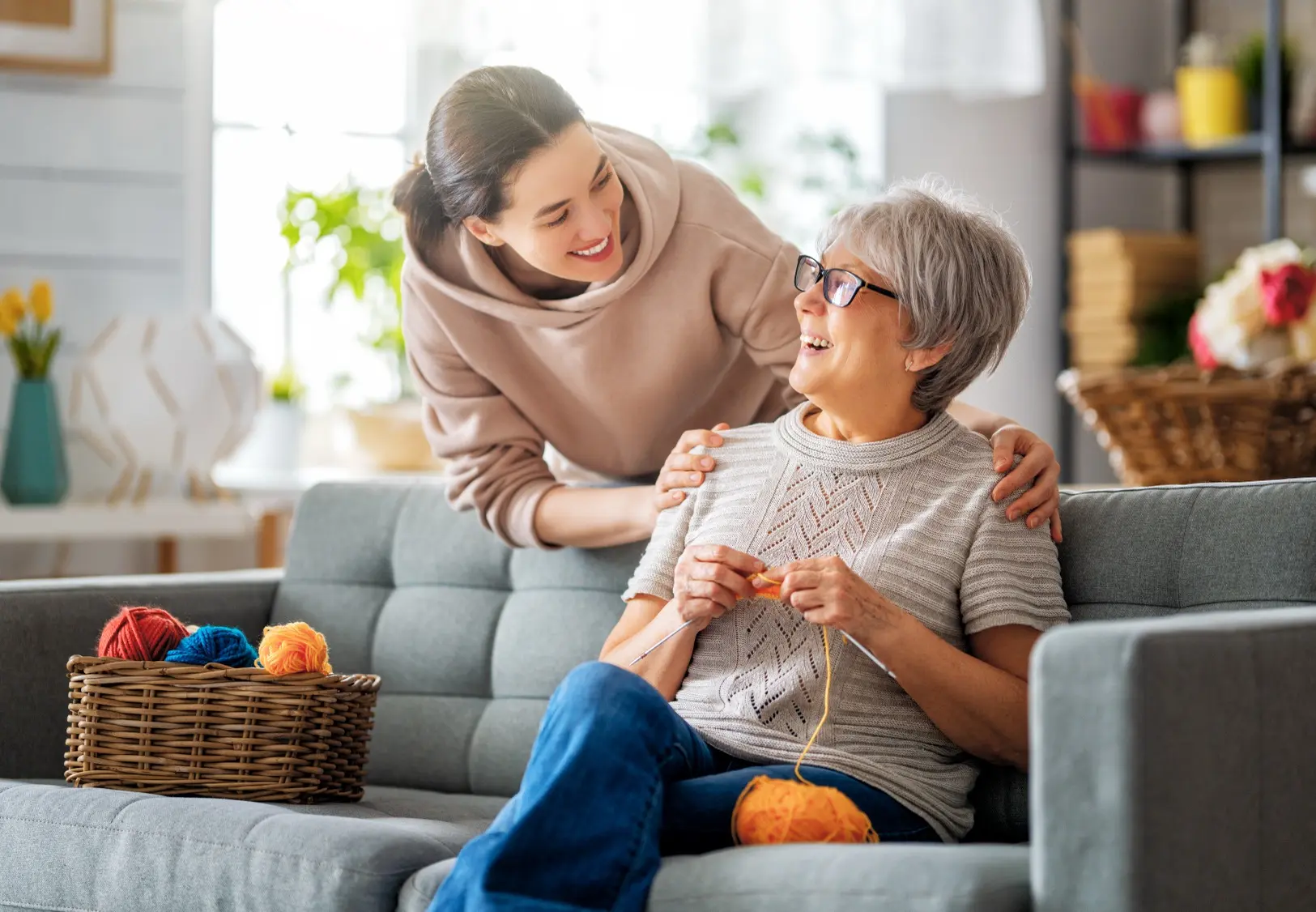 Ältere Frau mit grauen Haaren und Brille sitzt auf einem Sofa und strickt mit orangem Garn, jüngere Frau mit braunem Pullover steht hinter dem Sofa und legt die Hände liebevoll auf die Schultern der älteren Frau, beide lächeln sich an, im Hintergrund heller Wohnraum mit Pflanzen und Regalen.